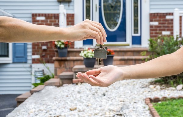 Person handing house key to another person in front of new home.